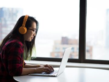 Asian woman with headphones using laptop for remote work in a city setting.