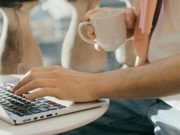 Close-up of a person working outdoors with a laptop and coffee in hand.